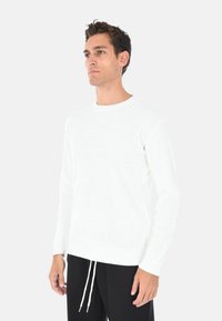 Maglione bianco a costine con maniche lunghe e scollatura rotonda. Presenta un tessuto testurizzato e polsi aderenti. Abbinato a pantaloni neri con coulisse.