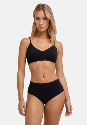 Femme aux longs cheveux blonds portant un bralette noir et un sous-vêtement noir taille haute, debout devant un fond clair uni.
