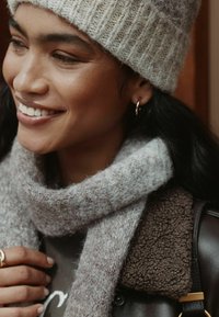 Knit gray-beige beanie, gray scarf with a soft texture, brown fluffy jacket collar, gold hoop earring, and a dark shirt underneath.