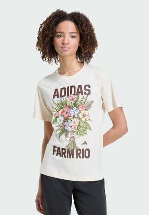 Giovane donna con capelli ricci che indossa una maglietta crema Adidas Farm Rio con una grafica colorata di bouquet floreale e pantaloni neri.