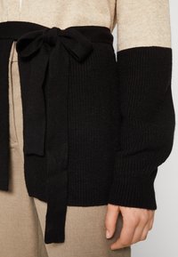 Svart stickad bälte knuten i en rosett på en beige och svart färgblockerad tröja, med beiga byxor och en avslappnad hand vid sidan.