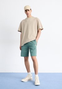 Camiseta oversized beige, pantalones cortos color teal con un pequeño logo y zapatillas blancas de caña alta. Estilo simple y relajado, presentado sobre un fondo liso.