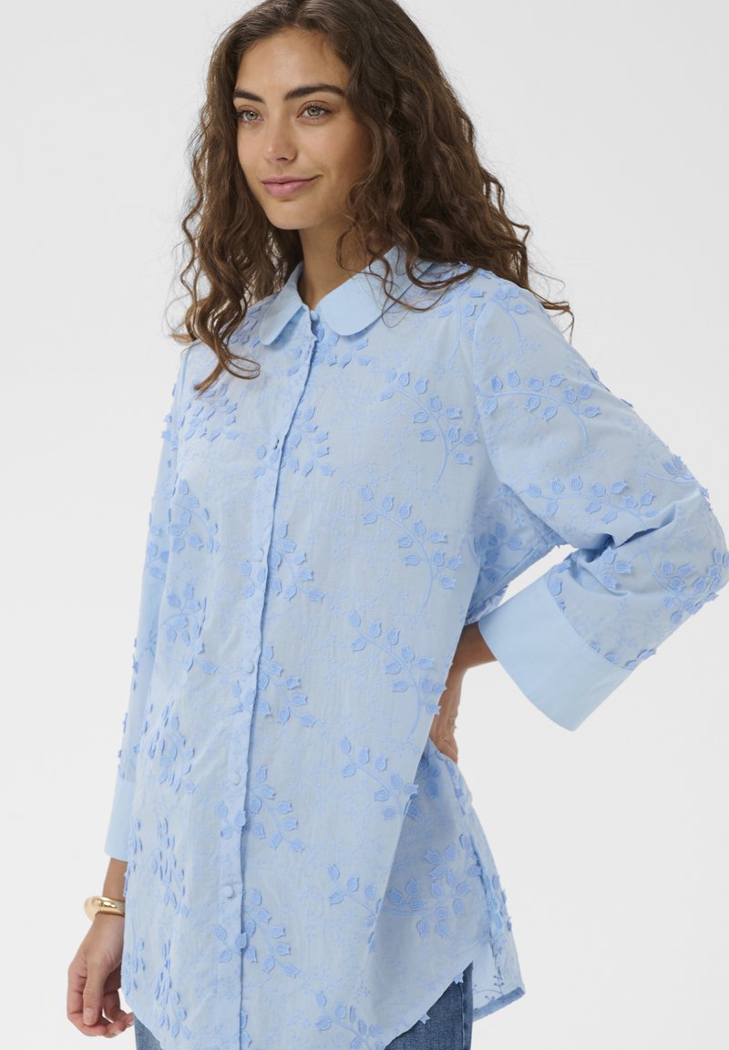 Naine pikad lokkis juuksed, kandes helesinist lillemustrilist tikanditega pika varrukaga särki, millel on nööbitavad mansetid ja krae, poseerimas käsi puusal.