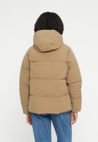 Giacca a vento marrone con cappuccio e motivo di trapuntatura orizzontale. Presenta una vestibilità comoda con maniche leggermente sovradimensionate.