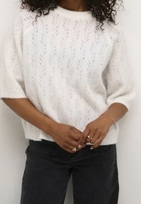 Pull en tricot blanc avec motif de feuilles, manches courtes et col rond, associé à un jean foncé. Tissu texturé et doux avec une coupe décontractée.