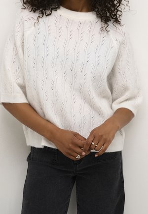 Pull en tricot blanc avec motif de feuilles, manches courtes et col rond, associé à un jean foncé. Tissu texturé et doux avec une coupe décontractée.