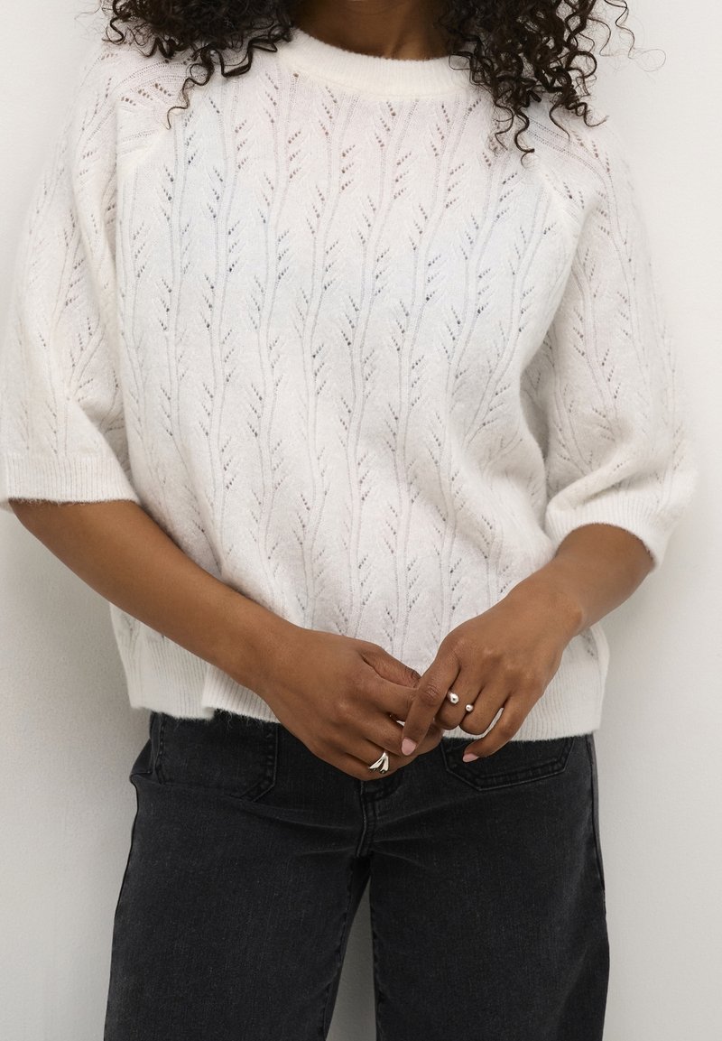 Pull en tricot blanc avec motif de feuilles, manches courtes et col rond, associé à un jean foncé. Tissu texturé et doux avec une coupe décontractée.