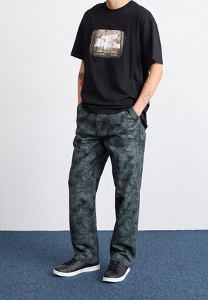 Personne portant un t-shirt graphique noir, un pantalon camouflage vert foncé et des baskets noires à semelles blanches, debout sur un tapis bleu.