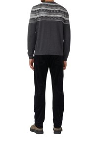 Homme portant un pull à motifs sombres, un pantalon noir et des chaussures brunes, se tenant de dos face à la caméra sur un fond blanc.