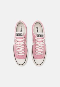 Rosa dukskor med vita snören, gummitå och svart sula. Har Converse-logga på tungan och stjärnaccenter på sidorna.