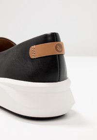 Chaussure noire en synthétique à enfiler avec une semelle en caoutchouc blanche, présentant un accent en cuir tan au talon et des détails de coutures subtils.