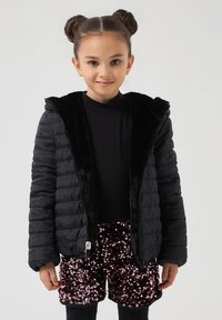 Giacca a sacco nera con colletto in pelliccia sintetica, chiusura con zip e design trapuntato; abbinata a pantaloni corti rosa lucidi con paillettes e un top nero.