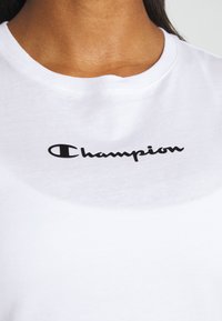 Gros plan du cou et du haut de la poitrine d'une personne portant un t-shirt blanc avec le logo noir "Champion" centré sous l'encolure.