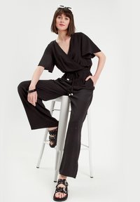 Schwarzer Overall mit weitem Bein, kurzen Ärmeln und verstellbarem Bund. Kombiniert mit schwarzen Sandalen, die mit silbernen Nieten und Schnallenverzierungen ausgestattet sind.