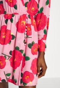 Florales Kleid aus pinkfarbenem Stoff mit großen roten Rosen, grünen Blättern und seitlichen Knopfverschlüssen. Lange Ärmel mit geraffter Taille.