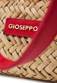 Sac en paille tissé avec une étiquette en cuir rouge portant des lettres dorées, mettant en avant un design complexe et une texture naturelle.