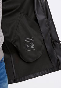 Svart vattentät och vindresistent jacka med slät textur, med en innerväska märkt "LINDBERGH" och skötselsymboler tryckta i vitt.