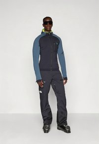 Navy and light blue athletic jacket with a zip, paired with dark pants featuring zippers. Model wears sunglasses and ski boots.