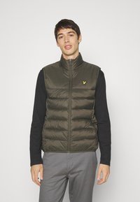 Zaļš pūkains veste ar augstu apkakli, ar priekšējo rāvējslēdzēju un mazu dzeltenu logo uz krūtīm, uzvilkta uz melnas garām piedurknēm krekla.