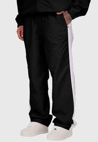 Melna sporta bikses ar baltām sānu svītrām un elastīgu jostu, ar teksturētu logotipu. Apvienotas ar baltām sporta apaviem.