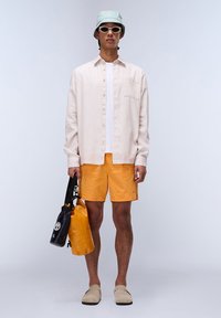 Light pink long-sleeve shirt, white t-shirt, orange shorts, beige slip-on shoes, black and orange bag, mint green bucket hat, white sunglasses.