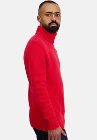 Pull à col roulé rouge en tricot avec poignets et ourlet côtelés, présentant une coupe décontractée et une texture lisse. Porté avec un jean bleu foncé.