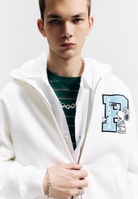 Witte zip-up hoodie met een blauw-zwart "Peanuts"-ontwerp, gedragen over een groen gestreept shirt, gecombineerd met een zilveren armband met ketting.