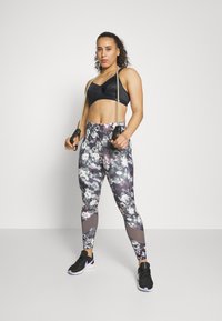 Svart sportsbh, høy midje leggings med blomstermønster i grått og lilla, holder på motstandsbånd, iført svarte treningssko.
