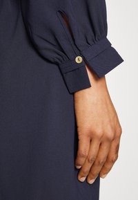 Blusa azul marinho com mangas de tecido franzido, fecho de botão e fenda lateral; textura suave e corte ajustado.