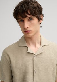 Beige Kurzarmhemd aus strukturiertem Stoff mit einem Kragen und zwei Knöpfen. Enganliegende Form, lässiges Design.
