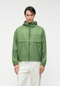 Ανοιχτό πράσινο αδιάβροχο μπουφάν με κουκούλα, φερμουάρ και μπροστινές τσέπες. Διαθέτει οριζόντια ραφή στο ύψος του στήθους και λογότυπο.