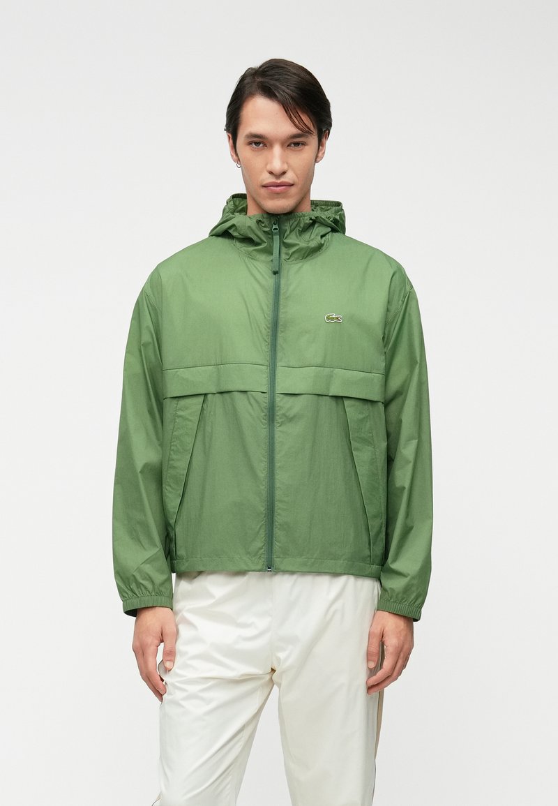 Ανοιχτό πράσινο αδιάβροχο μπουφάν με κουκούλα, φερμουάρ και μπροστινές τσέπες. Διαθέτει οριζόντια ραφή στο ύψος του στήθους και λογότυπο.