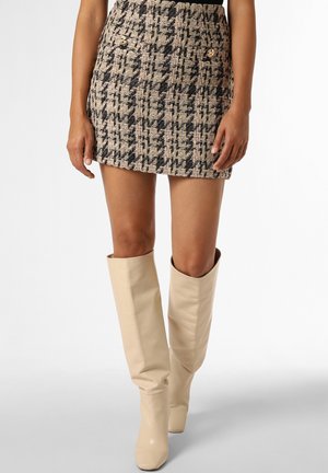 Femme portant une mini-jupe en tweed beige et noire avec des boutons dorés, associée à des bottes en cuir crème montant au genou.