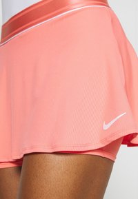 Falda deportiva rosa con textura acanalada, banda de acento blanca en la cintura, con un pequeño logo blanco de Nike en el dobladillo.