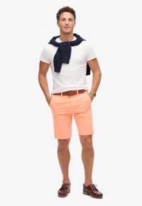 Pantaloni corti di un vivace color corallo, in tessuto di cotone, con una vestibilità regolare. Indossati con una t-shirt bianca a collo rotondo e un maglione blu navy appoggiato sulle spalle. Cintura in pelle marrone.