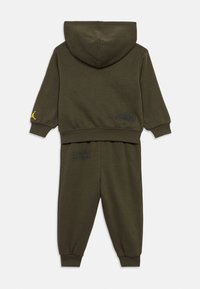 Ελαιοπράσινη φόρμα με κουκούλα, φτιαγμένη από μαλακό ύφασμα. Διαθέτει κίτρινο λογότυπο στο μανίκι και μαύρες εκτυπώσεις στο μπουφάν και τα παντελόνια.