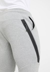 Närbild av ljusgråa sweatshirtbyxor med en svart lodrät dragkedjeficka på låret, bärs av en person i vit skjorta.