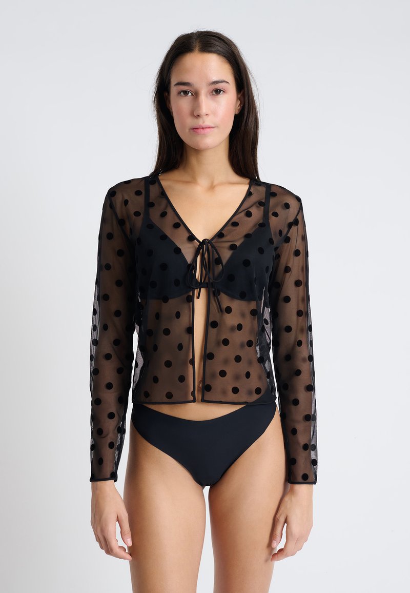 Top de malla negro con lunares, de manga larga y cierre tipo lazo, con escote en V profundo y textura transparente, superpuesto sobre un bikini negro.