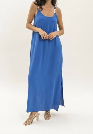 Femme portant une robe maxi bleue sans manches avec des bretelles à boucle dorée et des sandales à talons hauts dorées à lanières, debout sur un fond clair uni.
