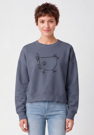Junge Person mit kurzen braunen Haaren, die einen grauen Sweatshirt mit einer einfachen schwarzen Strichzeichnung eines Huhns und hellblaue Jeans trägt.