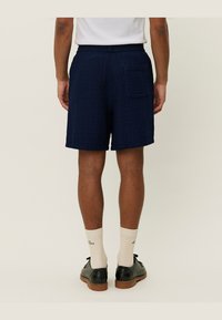 Pantaloni scurți de culoare albastru marin, confecționați dintr-un material texturat, cu un elastic la brâu, având un buzunar pe spate și o croială lejeră. Purtați cu șosete deschise la culoare și pantofi negri.