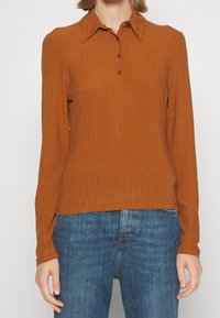 Long-sleeve ribbed top in warm brown, featuring a collar and three buttons. Made from a textured fabric, paired with denim jeans.