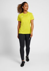 Lys gul træningstrøje med korte ærmer, parret med tætsiddende sorte leggings med logo og detaljer. Modellen står i en neutral setting.