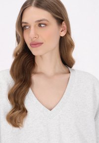 Pull V-col à col gris clair avec bordures côtelées, coupe ample, fabriqué en tissu doux. Le modèle a des cheveux bruns ondulés encadrant le visage.