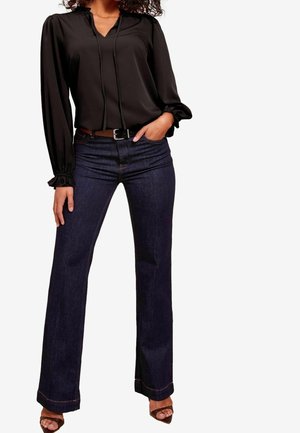 Vrouw die een zwart blouse met lange mouwen en ruches aan de manchetten draagt, donkerblauwe bootcut-jeans, een bruine riem en bruine open teen hakken, met haar hand in haar zak.