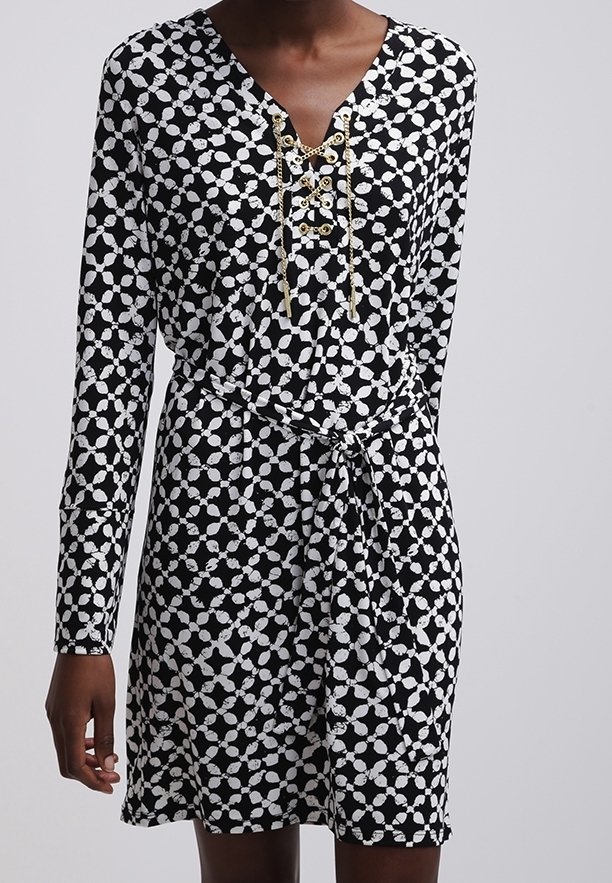 Robe à motifs noir et blanc avec manches longues, détail chaîne dorée lacée au niveau de l'encolure, et ceinture en tissu nouée à la taille.
