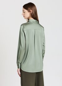 Donna con capelli castani lisci che indossa una blusa di satin verde chiaro larga e pantaloni verde oliva scuro, rivolta di spalle contro uno sfondo semplice.