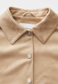 Camicia in suede beige con colletto, bottoni a pressione argento sulla parte anteriore e una finitura morbida e testurizzata. Anello per appendere visibile sul collo.