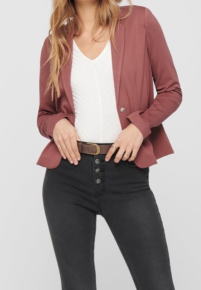 Femme portant un blazer mauve sur un haut blanc en V avec un jean noir à taille haute et une ceinture marron, mains ajustant la ceinture.
