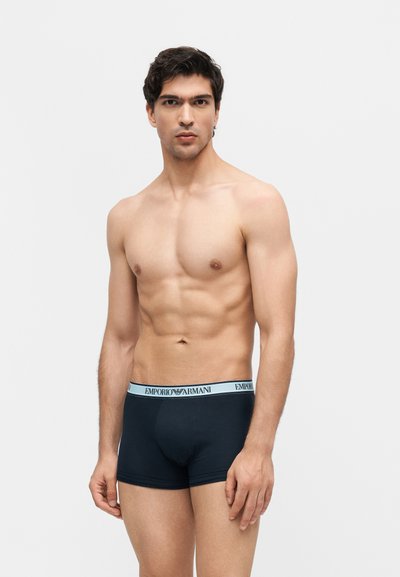 Φορώντας μαύρα εσώρουχα Emporio Armani, ένας γυμνασμένος νεαρός άνδρας με σκούρα μαλλιά στέκεται μπροστά σε λευκό φόντο.